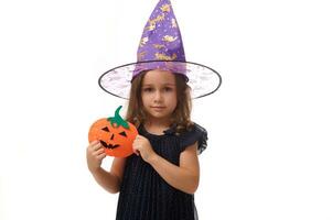 Isolated portrait on white background with copy space of beautiful baby little girl, 4 years old pretty kid wearing a wizard hat, holding a homemade felt-cut pumpkin, symbol of the Halloween party photo