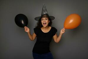 Happy joyful beautiful Hispanic woman wearing a wizard hat, holds two air balls of orange and black color, looks at camera posing against a gray background with copy space. Halloween concept photo