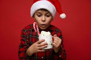 Close-up portrait of an amazed boy looking with surprise at a mug with hot chocolate drink decorate with marshmallows, sugary candy cane lollipop. Christmas concept on red background with space for ad photo