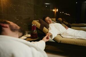 Newlywed, beautiful couple of an attractive woman in white bathrobe lying on sun lounger and looking at her loving husband holding her hand in his hand, enjoying their honeymoon at wellness spa resort photo