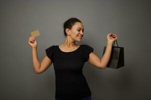 Pretty smiling African woman dressed in black holds a shopping bag and gold credit card, shows them posing to camera, isolated over gray wall background with copy space for Black Friday advertisement photo
