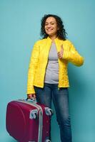 Beautiful delighted curly-haired woman in bright yellow jacket, holding her suitcase and showing thumb up, smiling toothy smile looking at camera, isolated over blue background with copy ad space photo