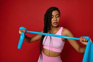 Determined athlete, African sportswoman in pink sportswear holding an elastic fitness strap and stretching arms, isolated over red background with copy space. Body positivity, fitness, sport concept photo