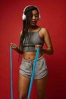 Young athletic African woman with Afro pigtails wearing wireless headphones and gray top and shorts, exercising with fitness elastic strap against red colored background with copy space photo