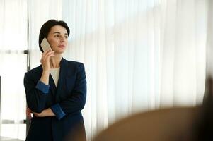 Beautiful woman in business casual suit talking on a mobile phone, standing by the window in a hotel room, looking thoughtfully from it into the distance at the cityscape during her business trip photo