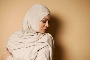 Close-up of serene elegant Muslim woman in traditional formal dress looking down on beige background with copy space photo