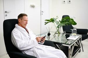 Relaxed handsome mature 50 years old businessman in white terry bathrobe sitting on an armchair with a smart phone and browsing, writing text messages at wellness spa center photo