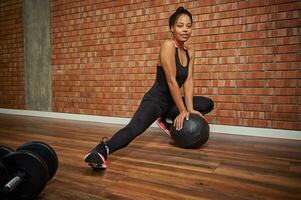 Charming young African American female athlete in a stylish tight-fitting tracksuit performing side lunges, leaning her hands on a medicine ball during sports training workout photo