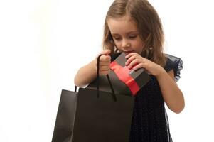 Concept for Black Friday, isolated portrait of a baby girl in dark blue evening dress putting a gift with red ribbon into a black packet, copy space. Shopping, sales and purchases concept. photo