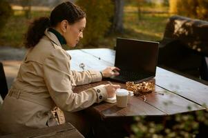 africano americano mujer en beige casual Saco se sienta en un de madera banco en un café y trabajos de forma remota, toma notas en un ordenador portátil y tipos en un ordenador portátil teclado. ventajas de distante trabajo y en línea estudiando foto