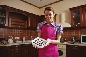 Attractive young confectioner wearing pink apron, standing in the middle of her home kitchen with a box of handmade chocolate truffles photo