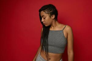 Side portrait of confident African woman with fit body in gray sportswear with stylish dreadlocks posing looking down on red background with copy space photo