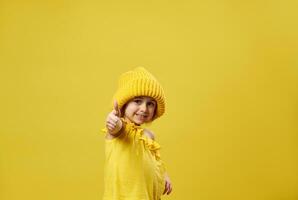 Beautiful little girl wearing yellow woolen hat shows thumb up to the camera and smiles while posing on a yellow background with copy space photo