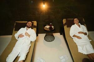 Relaxed happy loving young married couple smiles toothy smile looking at camera while resting in white terry bathrobes lying down on chaise lounges in lounge area of a luxury wellness spa complex photo