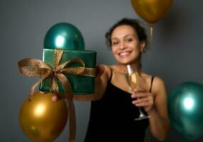 Focus on a Christmas gift in the hand of pretty woman in black evening dress, holding a flute of champagne and cutely smiling looking at camera, over gray background with golden and green air balloons photo