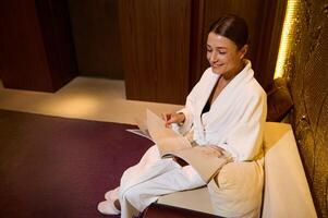 Overhead view of a middle aged elegant attractive European woman, dressed in white terry bathrobe, smiles toothy smile, relaxing, reading magazine while resting at wellness spa lounge area photo