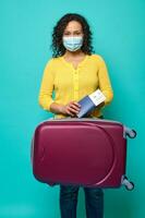 Young woman in protective medical mask, wearing bright yellow sweater posing with suitcase, passport and boarding pass in her hands. Travel, air flight journey concept on blue background, copy space photo