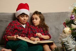 Beautiful curious children reading book, fairy tales while sitting on the couch near a Christmas tree in the living room photo