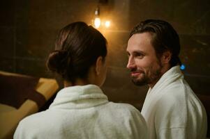 Close-up of a Caucasian middle aged newlywed, married couple in bathrobes sitting face to face and looking at each other, relaxing in luxury health spa resort photo