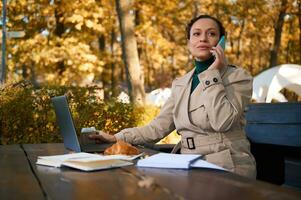 Confident business woman looking to the side, talking on mobile phone, sitting on a wooden bench in front of laptop and enjoying coffee break in a cozy cafe against golden autumn oak grove background photo