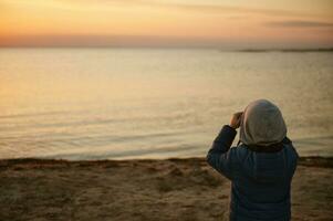 curioso pequeño chico mirando a el hermosa costero naturaleza a el puesta de sol mediante el binocular foto