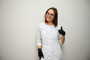 Beautiful young brunette woman, medic intern, female doctor in white uniform showing thumb up against white wall background with copy space photo