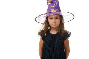 Portrait of beautiful confident little girl wearing a wizard hat and dressed in stylish carnival dress, looking at camera posing with crossed arms against white background, copy space. Halloween photo