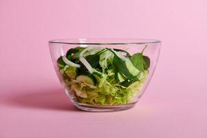 Close up of green salad with herbs, lettuce, cabbage and cucumber in transparent glass bowl on pink background photo