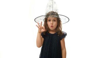 Isolated waist length portrait of pretty little witch girl wearing a wizard hat and dressed in stylish carnival dress, gesturing, showing OK sign with fingers. Halloween concept on white background photo