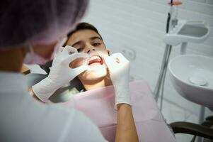 Overhead view of pediatric dentist putting cheek dental retractor on child patient before oral treatment. School boy at the dentist's appointment. Dental caries treatment and healthy teeth concept. photo