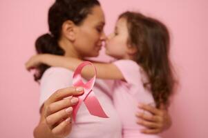 Soft focus on pink satin ribbon, symbol of International Breast Cancer Awareness Day, against blurred background of loving mother and lovely daughter. Female healthcare and medical education concept photo