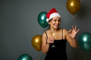 bonito mujer con hermosa sonrisa en Papa Noel sombrero participación un champán flauta y demostración un Okay firmar en contra brillante oro y verde aire globos en gris antecedentes con Copiar espacio para Navidad anuncio foto