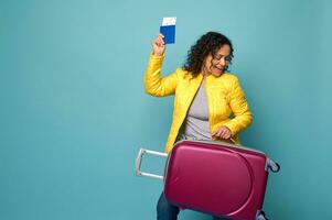 Funny joyful woman traveler tourist with curly hair, dressed in yellow jacket raised her arms with passport and ticket and dances with a luggage in her hands, enjoying her vacation. Travel concept photo