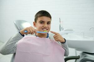 hermoso colegio chico se sienta en del dentista silla, mira a cámara participación cepillo de dientes cerca su boca y puntos con dedo en su hermosa con dientes sonrisa con sano blanco dientes. oral higiene concepto foto