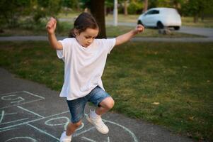 de cerca de pequeño caucásico niña jugando rayuela en asfalto. niño jugando rayuela juego en patio de recreo al aire libre en un soleado día. calle para niños juegos en clásicos foto