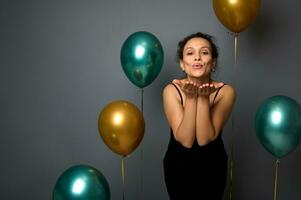 Attractive Hispanic woman dressed in elegant black dress stands against gray background with golden green metallic inflated balloons, send air kisses looking at camera. Christmas celebration concept photo