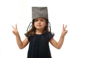 Portrait of beautiful Caucasian European girl having fun, putting a black shopping packet on her head, looking at camera, showing peace sign with her fingers. Black Friday concept photo