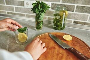 Close-up of a hand tossing a slice of lemon and mint leaves into a transparent glass. Sliced lemon, knife on a wooden board near a transparent glass with mint on the kitchen countertop photo
