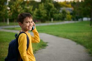 adorable colegial vistiendo amarillo camisa de entrenamiento con mochila hablando en móvil teléfono en público parque, yendo hogar después escuela, , sonriente con con dientes sonrisa a el cámara foto