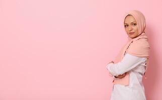 Confident portrait of Arabic muslim beautiful woman with attractive look and gaze, wearing a pink hijab and standing sideways against colored background with copy space photo