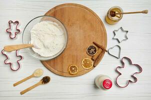 Flat lay of ingredients for preparing dough and gingerbread cookies, cutting molds, bottle with plant based milk, glass jar with honey and a bowl with flour on a round wooden board photo