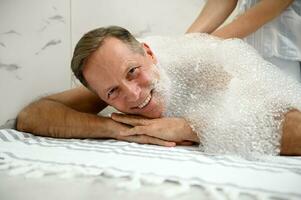 Close-up of a handsome mature European man smiling with beautiful toothy smile, looking at camera while relaxing in Turkish hammam with soap foam on his back photo