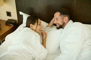 Young handsome European man, husband smiles toothy smile looking tenderly at his beloved wife, lying next to her in bed after waking up. Loving couple enjoying honeymoon together. Top view photo