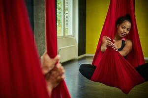 Reflection on the mirror of a sporty African pretty woman with slim body hanging on a red hammock, relaxing during aerial fly yoga stretching exercises photo