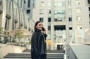 Cheerful young brunette woman talking on mobile phone smiling toothy smile looking over shoulder at camera climbing stairs to modern office corporate high-rise building. photo