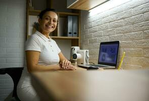 African American pretty female doctor smiles looking at camera sitting in front of a laptop with panoramic X-ray photograph and contemporary portable X-ray machine in dentists office photo