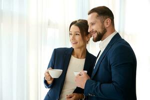 Two young successful handsome business partners standing next to each other with a cup of coffee in their hands and smiling, confidently looking out the window. Copy space for ad photo