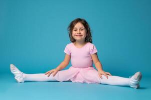 hermosa pequeño niña bailarina practicas extensión su piernas y linda sonrisas a el cámara, aislado en azul antecedentes foto