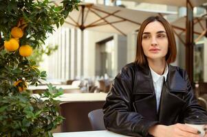 Focus on a beautiful young short haired brunette woman looking away sitting at a table with a takeaway paper cup of hot drink on the summer terrace of a cozy cafe with a tangerine decorative tree photo