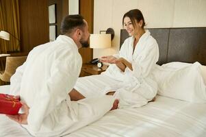 Handsome romantic husband, man hiding gift box behind his back, surprising his beloved girlfriend, wife on special day, resting together in bed. Valentine's day, birthday, anniversary, wedding date. photo